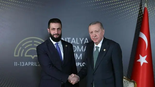 صورة للرئيس التركي رجب طيب إردوغان (يمين) يصافح نظيره السوري أحمد الشرع، ويظهر في يمين الصورة العلم التركي ذو اللون الأحمر الذي يتوسطه هلال ونجم باللون الأبيض، وكتب على الجدار الذي يقفان أمامه Antalya Diplomacy Forum، 11-13 April 2025 (منتدى أنطاليا للدبلوماسية، 11-13 أبريل/نيسان 2025)
