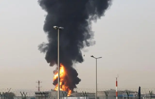 تصاعد الدخان واللهب من منطقة قرب مطار دبي الدولي في دبي، بالإمارات، ويمكن رؤية بعض المباني المنخفضة، محاطة بسياج أمني يبدو أنه مصنوع من أسلاك شائكة أو أسلاك حادة.
