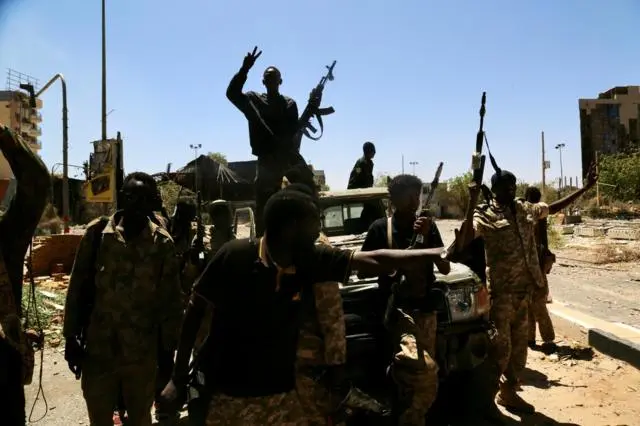 صورة توضح مجموعة من القوات التي تدعم الجيش السوداني، مع وجود ضابط جيش يقف بينهم حاملاً بندقية